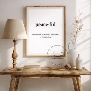May include: A framed print displaying the word "peaceful" and its definition. A wooden table supports the framed print, a lamp with a beige lampshade, and decorative vases. The style is minimalist and calming, with natural wood tones.