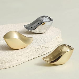 May include: Three abstract bird-shaped decorative objects. One is gold, one is silver, and the third is also gold. The objects have a smooth, polished finish and are arranged on a textured, off-white surface. The design is modern and minimalist.