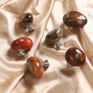 Może przedstawiać: Kolekcja dekoracyjnych gałek do szafek. Gałki wykonane są z polerowanego kamienia w odcieniach brązu, czerwieni i kremu, z unikalnymi wzorami. Każda gałka ma srebrną metalową podstawę.