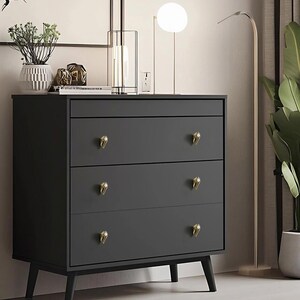 May include: A dark gray three-drawer dresser with gold-colored, decorative drawer pulls. The dresser has a minimalist design with tapered legs. A modern lamp and decorative items are on top of the dresser.