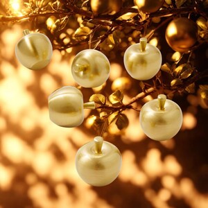 Op de afbeelding: Verschillende gouden, appelvormige ornamenten hangen aan een tak. De ornamenten hebben een glad, reflecterend oppervlak en een kleine steel. De achtergrond is een vage, warm getinte scène, die een feestelijk thema suggereert.