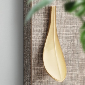 Puede incluir: Un tirador de armario dorado en forma de hoja. El tirador está montado sobre una superficie de madera texturizada gris-marrón. El diseño es elegante y moderno, con una forma suave y curvada. El tirador es un elemento de ferretería decorativo.