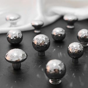 以下が含まれることがあります： ファセット加工とハンマー加工が施された、銀色のキャビネットノブのコレクション。丸型やキノコ型など、さまざまな形状のノブが暗い表面に配置されています。ノブはおそらく金属製で、光を反射しています。
