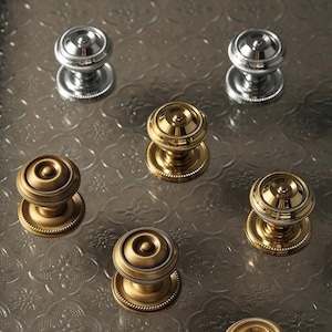 以下が含まれることがあります： シルバーとゴールドの色合いの装飾的なドアノブの詰め合わせ。ノブは、丸いベースと詳細な丸みを帯びた上部を備えたクラシックなデザインです。ノブは、テクスチャ加工された透明な表面に配置されています。