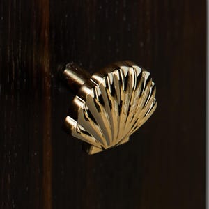 Może przedstawiać: Zbliżenie złotego uchwytu do szafki w kształcie muszli. Uchwyt jest zamontowany na ciemnobrązowej drewnianej powierzchni. Muszla ma szczegółowe żebra i gładkie, wypolerowane wykończenie. Ten dekoracyjny element dodaje meblom nadmorskiego charakteru.