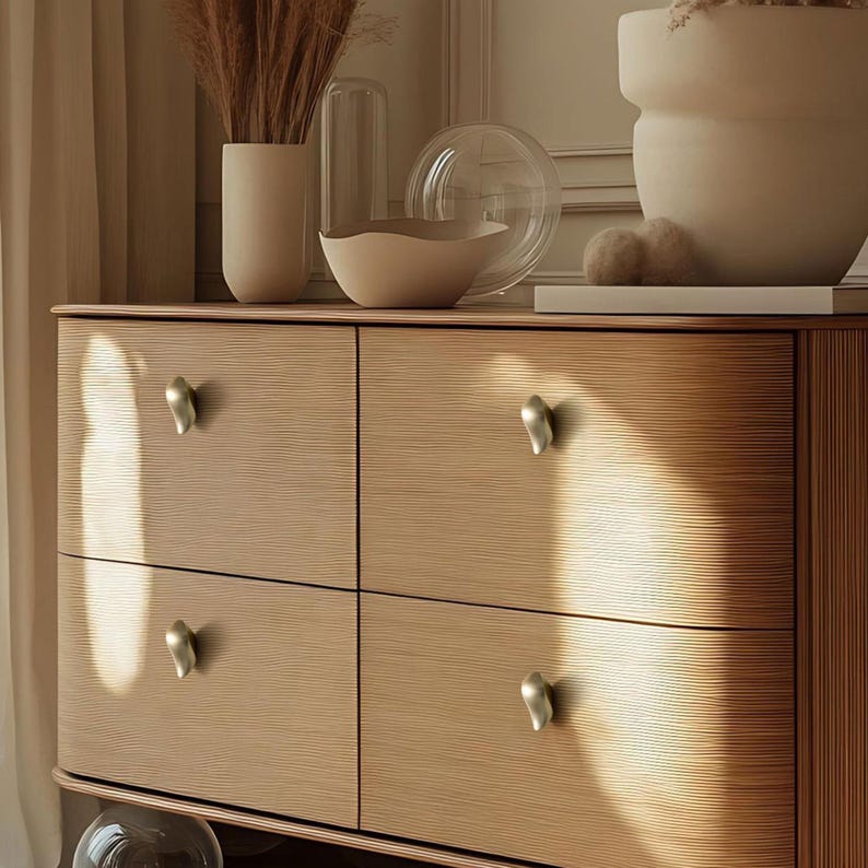 May include: A light wood dresser with four drawers, each with a unique silver-toned knob. The dresser is topped with decorative vases and a glass bowl. The wood grain is visible, and the overall aesthetic is modern and minimalist.