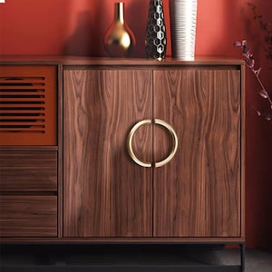 Könnte beinhalten: Ein Holzschrank mit warmbraunem Finish und einem goldfarbenen Griff. Der Schrank hat zwei Türen und eine Schublade. Der Schrank ist Teil einer größeren Möbelgarnitur, mit einer roten Wand im Hintergrund.