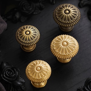 Könnte beinhalten: Vier antike Messing-Schrankknöpfe mit floralem Design. Die Knöpfe haben eine runde Form mit einem detaillierten Blütenblattmuster und einem Perlenrand. Sie sind vor einem dunklen Hintergrund platziert.