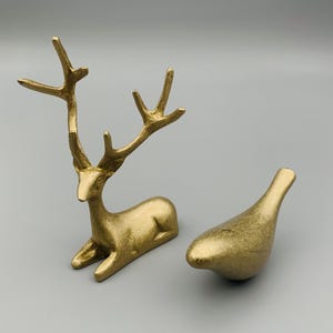 Puede incluir: Dos figuras decorativas doradas. Una es un reno estilizado en posición de descanso, con grandes cuernos. La otra es una pequeña escultura redondeada con forma de pájaro. Ambas tienen una superficie texturizada y están sobre un fondo gris.