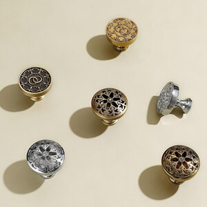 May include: A collection of decorative cabinet knobs in various designs and finishes. The knobs feature intricate patterns, including floral and geometric motifs, in gold, silver, and bronze tones. Some knobs have a solid design, while others have openwork detailing.