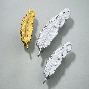 Op de afbeelding: Drie decoratieve, veervormige kastknoppen in goud en zilver. De metalen knoppen hebben een gedetailleerd veermotief. Deze decoratieve handgrepen zijn geschikt voor meubels. Ze zijn ongeveer 10 cm lang.
