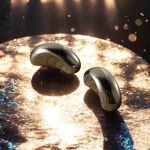 May include: Two polished, metallic, bean-shaped decorative objects. The objects have a smooth, reflective surface, and are positioned on a textured, golden-toned surface. The lighting creates highlights and shadows, enhancing the objects' form.