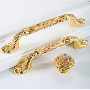 May include: Two ornate gold cabinet handles and a matching knob. The handles feature a filigree design and are curved with decorative ends. The knob is round and covered in small, clear crystals. The hardware is likely for furniture or cabinetry.