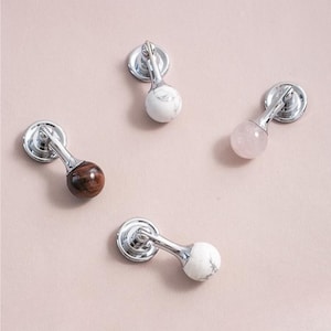 Op de afbeelding: Vier decoratieve kastknoppen met zilverkleurige hardware en bolvormige knoppen. De knoppen zijn gemaakt van verschillende materialen: wit marmer, een roodbruine steen, een roze steen en wit marmer met grijze adering.