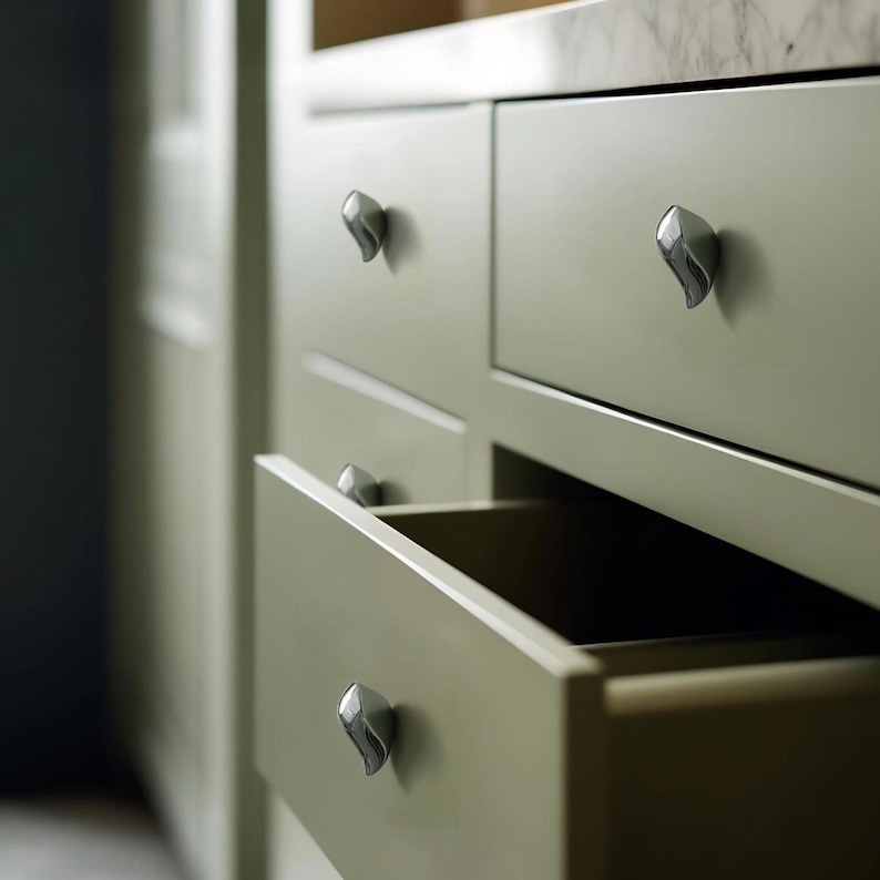 May include: Close-up of a sage green cabinet with multiple drawers. Each drawer features a silver, leaf-shaped handle. One drawer is open, revealing the dark interior. The cabinet is part of a larger kitchen or storage unit.