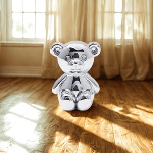 May include: A shiny, silver-colored teddy bear figurine. The bear is sitting with its arms at its sides. The bear has a smooth, reflective surface, and is sitting on a wooden floor. The background includes a window and curtains.
