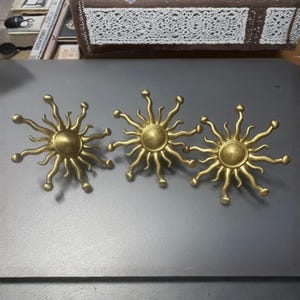 Könnte beinhalten: Drei goldfarbene, sonnenförmige Dekorationsgegenstände. Jede Sonne hat eine zentrale Scheibe mit Strahlen, die in kleinen Kugeln enden. Die Objekte sind in einer Reihe auf einer grauen Oberfläche angeordnet.