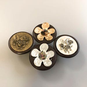 May include: Four decorative buttons with dark brown bases. Two buttons feature floral designs, one in gold and one in silver. The other two buttons have butterfly and rose designs, one in gold and one in silver. The buttons are arranged on a light gray surface.