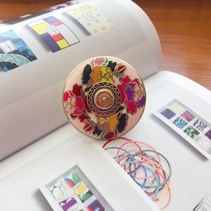 May include: A circular, pale pink decorative knob featuring a floral pattern in red, pink, yellow, and purple. The knob has a gold-coloured rim and centre detail. The knob is on a white surface with other design elements.