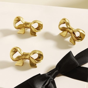 Puede incluir: Tres pomos de armario dorados con forma de lazo. Los pomos son de metal y tienen un acabado brillante. Una cinta negra está en la esquina inferior derecha. Los pomos están sobre una superficie blanca.