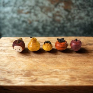 Op de afbeelding: Een verzameling van vijf houten fruitfiguren, waaronder een appel, een peer, een kaki en een vrucht met een lachend gezicht. De vruchten zijn gerangschikt op een houten oppervlak, waarbij hun natuurlijke kleuren en texturen worden getoond. De achtergrond is een gedempt groen.