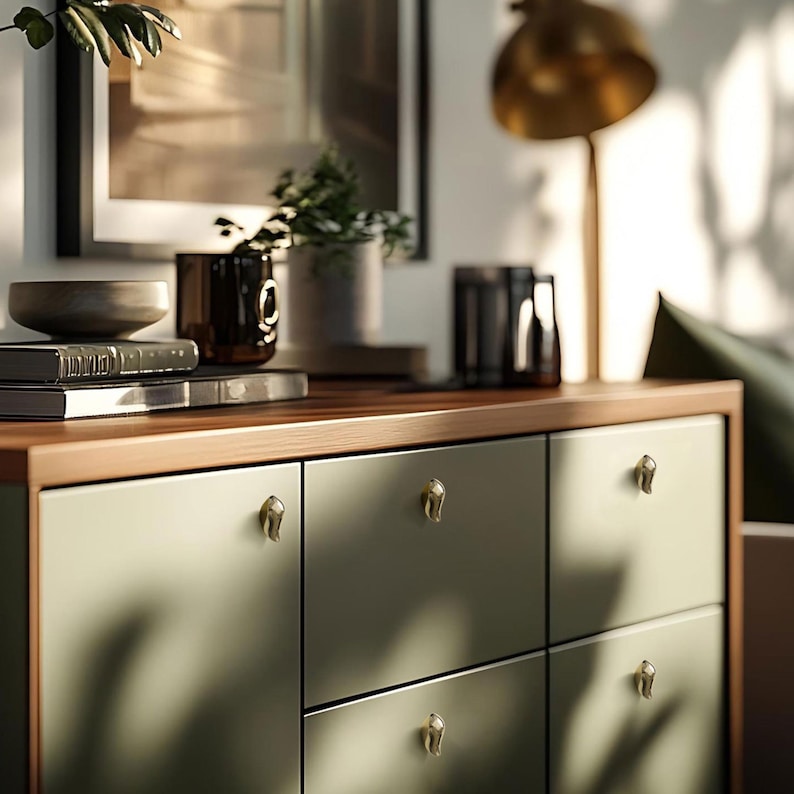 May include: A modern dresser with six drawers, painted in a muted green. The drawers have gold-colored, decorative pulls. The top of the dresser is a warm wood tone, and various decorative items are displayed on top, including books, a bowl, and a small plant.