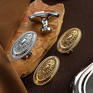 Könnte beinhalten: Eine Auswahl an dekorativen Schrankknöpfen und -griffen in Silber- und Goldtönen. Die Knöpfe haben eine ovale Form mit aufwendigen Details. Ein Knopf hat ein T-förmiges Design. Die Hardware wird auf einer Steinoberfläche präsentiert.