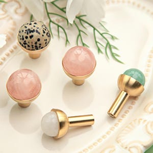 Può includere: Una collezione di pomelli decorativi per mobili con basi dorate e vari top in pietra. I pomelli presentano una pietra beige maculata, quarzo rosa, una pietra verde chiaro e una pietra bianca. Per mobili e decorazioni per la casa.
