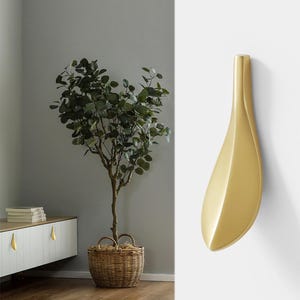 May include: A gold-colored cabinet pull in a leaf shape is mounted on a white wall. The pull has a smooth, curved design. In the background, a white cabinet with matching pulls and a potted tree with green leaves are visible.