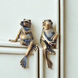 Puede incluir: Dos tiradores de armario de bronce con forma de rana. Cada rana está sentada, con una mano en la cara. Están montados en una puerta de armario blanca. Los tiradores tienen un aspecto envejecido y detallado.