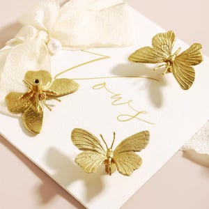 May include: Gold-coloured butterfly-shaped decorative hardware. The hardware is made of metal and has a textured surface. The butterflies are arranged on a white card with gold script. A white ribbon and pearl accent the card.