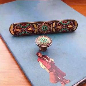 Könnte beinhalten: Dekoratives Hardware-Set mit einem langen Griff und einem runden Knauf. Beide Teile haben einen dunklen Hintergrund mit aufwendigen Mustern in Gold, Türkis und Rot. Das Hardware-Set liegt auf einer blauen Oberfläche.