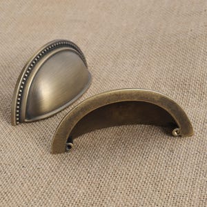 May include: Two antique brass-colored drawer pulls. One is a half-moon shape with a beaded edge, and the other is a simple curved handle. The hardware has a vintage aesthetic, suitable for furniture restoration or adding a classic touch to cabinets.