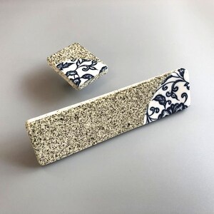 May include: Two ceramic cabinet pulls. One is rectangular, the other square. Both have a speckled, beige base and a corner section with a white background and a blue floral design. The pulls are set against a grey background.