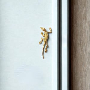 May include: A gold-colored lizard-shaped decorative accent is affixed to a white surface. The lizard is positioned as if climbing, with its legs and tail extended. The background includes a white frame and a section of a wood-grain surface.