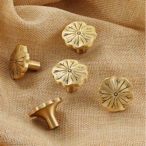 May include: Five brass cabinet knobs shaped like lotus flowers. The gold-coloured knobs feature a detailed leaf pattern and a short, cylindrical stem. They are arranged on a textured, tan fabric background.