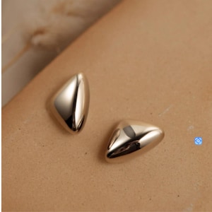 Könnte beinhalten: Zwei polierte, silberfarbene Ohrstecker. Jeder Ohrring hat eine glatte, abgerundete dreieckige Form mit einer reflektierenden Oberfläche. Die Ohrringe werden auf einer neutralen, hellbraunen Oberfläche präsentiert.