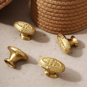 May include: Four gold-colored cabinet knobs with an oval, ornate design. The knobs have a detailed floral pattern and a rounded base. The knobs are arranged on a light gray surface, with a woven basket in the background.