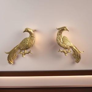 Puede incluir: Dos apliques de pared dorados con forma de pájaro. Cada pájaro está posado en una rama, con diseños detallados de plumas y alas. Los apliques están montados en una pared rosa claro, añadiendo un toque de elegancia.