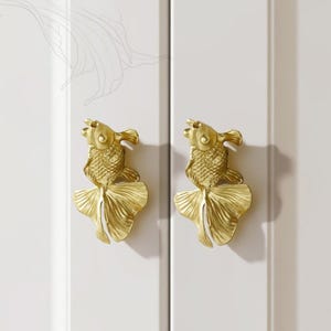 Puede incluir: Dos tiradores dorados con forma de pez. Los tiradores están montados en una puerta de armario blanca. Los peces tienen escamas detalladas y aletas caudales fluidas. Los herrajes del armario añaden un toque decorativo a los muebles.
