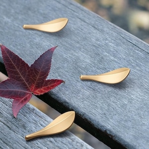 Puede incluir: Cuatro tiradores dorados en forma de hoja sobre una superficie de madera gris. Una hoja roja y granate está en primer plano. Los tiradores tienen un diseño suave y curvado, lo que sugiere un tema inspirado en la naturaleza.