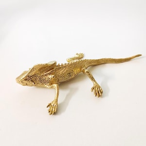 Puede incluir: Una figura de lagarto de metal de color dorado. El lagarto está tumbado con las patas extendidas y la cola curvada. La figura tiene detalles, incluyendo escamas y una superficie texturizada. Es un artículo decorativo.