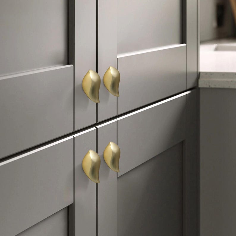 May include: Close-up of four gold leaf-shaped cabinet knobs on gray cabinet doors. The knobs are arranged in pairs, with two knobs on each set of doors. The cabinet doors are a neutral gray color, and the knobs provide a contrasting metallic accent.