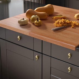 May include: A wooden countertop with various gourds and a knife. The gourds include butternut squash and small pumpkins. The countertop is above dark gray cabinets with gold-colored hardware. Diced squash is on the countertop.