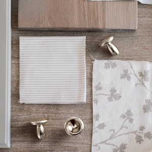 May include: An assortment of interior design components, featuring fabric samples with grey stripes and floral designs, a wooden plank, and three silver mushroom-shaped cabinet knobs. The items are displayed on a wooden surface.