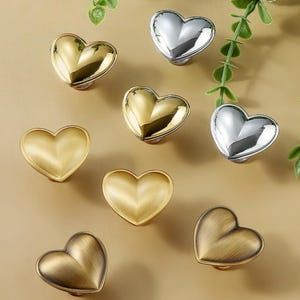 May include: A selection of heart-shaped cabinet knobs in gold, silver, and bronze. The knobs are displayed to show their different metallic finishes. Decorative hardware for furniture, ideal for drawers and cabinets.