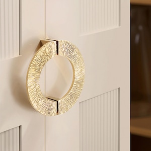 May include: A gold-coloured, textured ring pull handle mounted on a cream-coloured cabinet door. The circular ring has a rough, hammered appearance, and is attached to the door with a rectangular base.