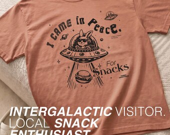 May include: A terracotta-colored short-sleeve t-shirt featuring a black graphic print of a rabbit wearing sunglasses inside a flying saucer. The text reads I Came in Peace. For Snacks. The design includes stars, a planet, and a burger being beamed up.