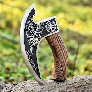 May include: A Viking-style axe with a curved blade and a wooden handle. The blade features a skull design and runes. The handle is made of dark wood. The axe is displayed outdoors against a blurred green background.