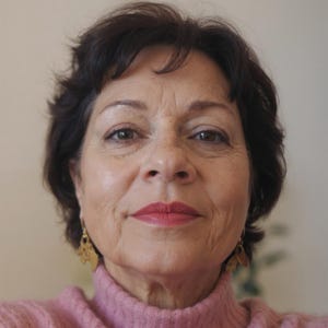 May include: A person with short dark brown hair wearing a pink ribbed turtleneck sweater and gold-toned dangling earrings. The earrings feature a cutout leaf or petal design. The person has a neutral expression with soft pink lipstick applied.